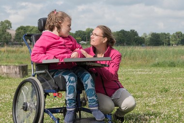 A woman kneels beside a girl in a wheelchair, and both are smiling at each other.